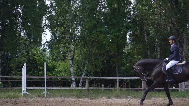 young girl riding a strong brown horse outdoors sand parkour dressage arena at dawn. Competitive training of riders. A woman in the festive uniform of a rider. sliding camera movement to the left. The