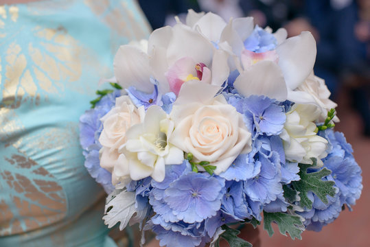Cropped shot of Caucasian young bridesmaid or female guest wearing a blue dress with silver details and holding a beautiful mixed bouquet featuring pale pink roses, white orchids and blue hydrangeas