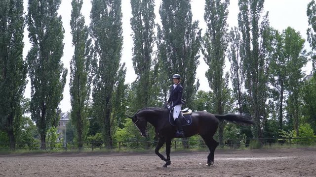 young girl riding a strong brown horse outdoors sand parkour dressage arena at dawn. Competitive training of riders. A woman in the festive uniform of a rider. sliding camera movement to the left. The