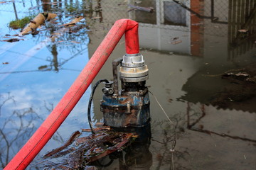 Partially submerged water pump pumping water from flooded garden through large red hose with house...