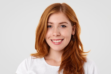 Studio shot of pleasant looking Caucasian female with gentle smile, freckled skin and make up, rejoices promotion at work, dressed in domestic clothes, isolated over white studio background.