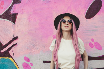 Happy stylish young hipster woman with long pink hair, hat and sunglasses on the street.