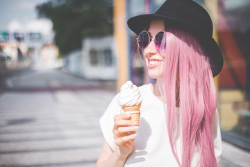 Happy young hipster woman with long pink hair, hat and sunglasses eating ice cream outdoors