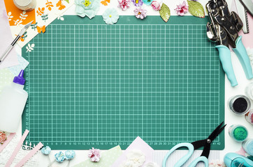 The cutting mat is surrounded by paper flowers, paper, tools and scrapbooking materials. Scrapbooking, top view, empty space in the center
