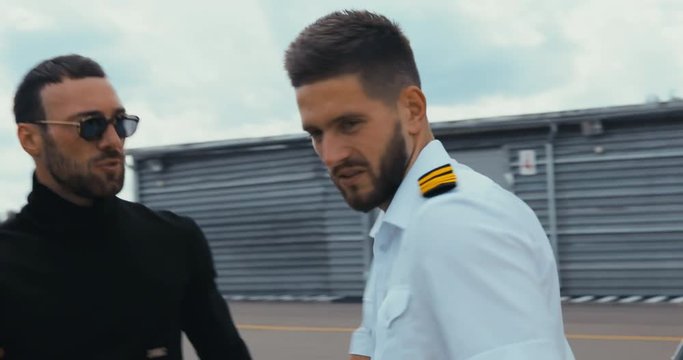 TRACKING commercial pilot in uniform greeting clients near small private helicopter on a landing point. 4K UHD