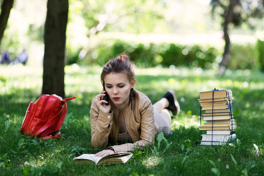 Young Girl Student Reading Book Lying On Grass In Park And Talking On Phone. Nearby There Are A Stack Of Books And A Red Backpack.