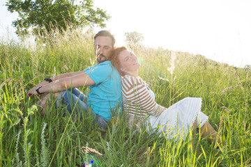 Portrait of carefree man and woman relaxing on the meadow with enjoyment. Man and woman sitting back to back each other and laughing while sitting on the grass with sunset light.