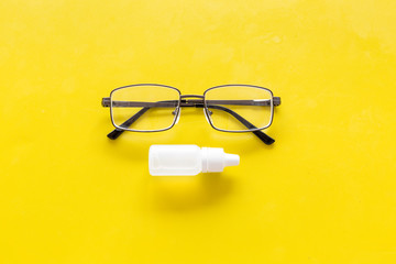 Eye drops in small bottle near glasses on yellow background top view copy space