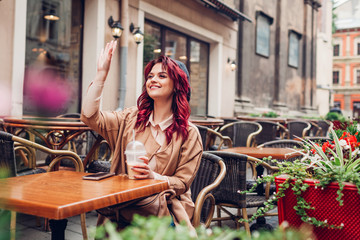 Obraz na płótnie Canvas Beautiful young woman waving at friends while having coffee in outdoor cafe. Meeting in the city