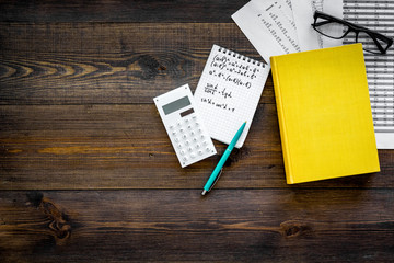 Math homework. Math textbook or tutorial near sheet with numbers, countes, calculator, notebook with formula on dark wooden background top view copy space
