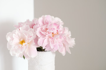 Angle of white and gray walls in room. On dresser stands white vase with pink flowers blooming peonies.