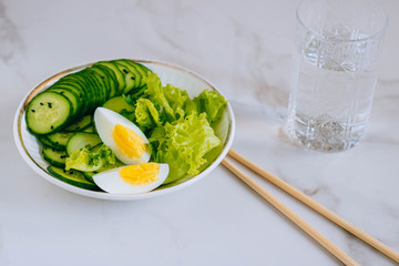 Healthy bowl, cucumber salad with eggs, coriander and glass of water on marble background