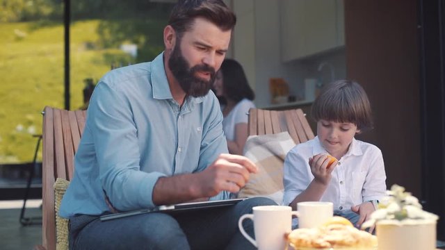 Father and little son sitting on terrace near beautiful modern house. Good-looking dad with beard giving laptop to kid. Caucasian family. Outdoors. Summertime. Vacation home.