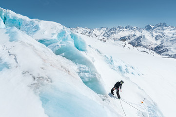 Naklejka na ściany i meble A professional mountaineer in a helmet and ski mask on insurance makes a nick-hole in the glacier against the backdrop of the Caucasian snow-capped mountains