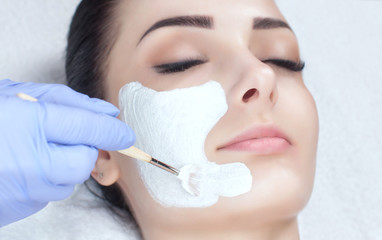 The cosmetologist for the procedure of cleansing and moisturizing the skin, applying a mask with stick to the face of a young woman in beauty salon. Cosmetology and professional skin care.