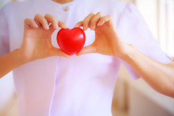 Fototapeta premium Red heart held by smiling female nurse's hand, representing giving effort high quality service mind to patient. Professional, Specialist in white uniform concept
