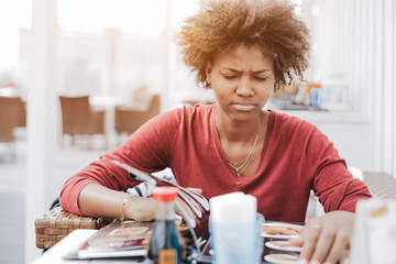 Cute curly African-American girl in street cafe is dissatisfied with an assortment of food in the menu; a young black female is angrily flipping through the menu pages while sitting in a restaurant