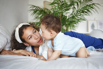 Obraz na płótnie Canvas People, maternity, childhood and family concept. Caring loving young mixed race mom lying on bed with her cute baby toddler, encouraging him while he is crawling, making first unbalanced movements