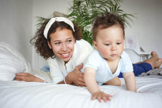 Indoor shot of stylish happy young Hispanic mother wearing white headscarf havig fun with adorable chubby 10 month old son, crawling on bed with serious look. Selective focus on woman's face