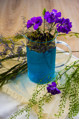 A glass brewer full of blue butterfly pea tea, with purple flowers and green branches next to it, on crumpled wrapping paper on a wooden table.