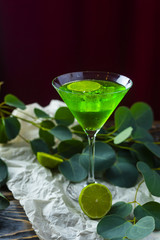A green drink with ice cubes in a glass for mojito on crumpled wrapping paper next to which lie slices of lime and a branch of green tropical leaves on a vintage wooden table.