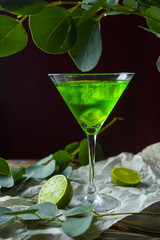 A green drink with ice cubes in a glass for mojito on crumpled wrapping paper next to which lie slices of lime and a branch of green tropical leaves on a vintage wooden table.