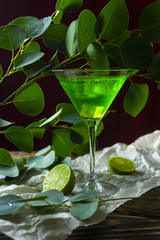 A green drink with ice cubes in a glass for mojito on crumpled wrapping paper next to which lie slices of lime and a branch of green tropical leaves on a vintage wooden table.