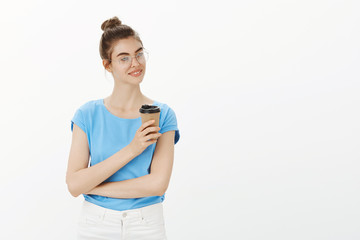 Great day to be productive. Good-looking feminine female in glasses and bun hairstyle, looking right with sensual smile, holding cup of coffee, having break during lunch at work over grey wall