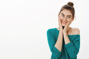 I am so happy to have such loving friends. Portrait of emotional pleased and tender female in loose sweater and glasses, holding palms on cheeks and smiling with cute expression, being touched