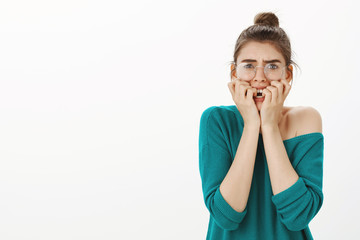 Studio shot of shocked and scared young woman in transparent glasses and loose sweater, frowning,...