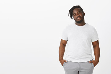 Portrait of easy-going plump african-american guy with braids, holding hands in pockets, tilting...