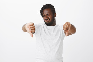 Portrait of disgusted or displeased african-american businessman in white t-shirt, showing thumbs down, frowning and staring with unimpressed disliking expression at camera, rejecting bad idea