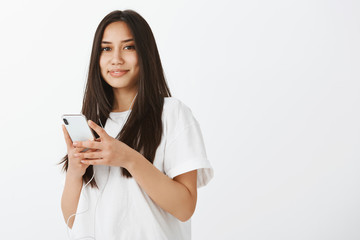 Waist-up shot of good-looking confident female coworker in white t-shirt, holding smartphone, listening music in earphones, smiling with tender and caring expression, enjoying nice songs