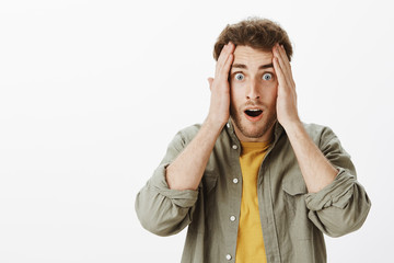 Guy being shocked, cannot understand how it happened. Portrait of stunned and speechless good-looking male with beard and curly hair, holding palms on face and dropping jaw from amazement