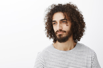 Fototapeta premium Waist-up portrait of handsome masculine dreamy male designer with curly haircut and stylish striped t-shirt, looking away and smiling sensually, recalling nice memories from past over gray wall