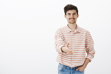 Neat polite man greeting new employer. Portrait of good-looking confident friendly male model with moustache and beard, pulling hand towards camera in handshake, welcoming newcomer over gray wall