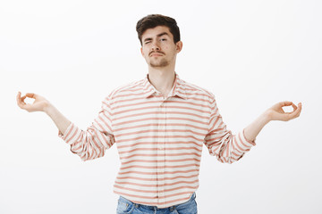 Fototapeta premium Studio shot of unfocused handsome male with moustache, spread hands in zen gesture and peeking with one eye, being calm and relaxed during meditation and yoga over gray background