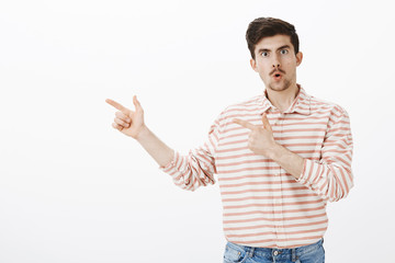 Wow, check this out. Indoor shot of amazed wondered man with moustache, folding lips and pointing at upper left corner from surprise and astonishment, standing over gray wall, discussing shocking news