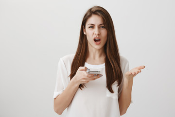 Who can make such stupid mistakes in spelling. Portrait of angry and annoyed attractive female businesswoman holding smartphone and shaking palm with displeased expression, showing her anger