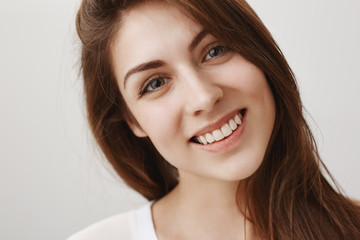 Fototapeta premium Close-up shot of charmed good-looking caucasian girl tilting head from interest and curiosity, showing pleasure to talk with speaker, smiling broadly at camera, gazing with admiration over gray wall