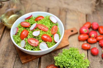 View from above of green salad Lollo Biondo with tomatoes and radishes in a white bowl on wooden table, a pitcher of oil in the background