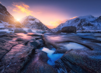 Colorful sunset in Utakleiv beach, Lofoten islands, Norway. Amazing scene with snowy mountains, stones, beautiful reflection in water, red sky, clouds. Winter landscape. Coast with stones in water