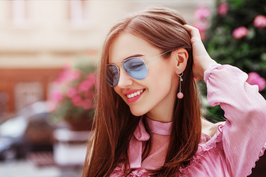 Outdoor close up portrait of young beautiful happy smiling woman wearing blue aviator sunglasses, trendy earrings, posing in street of european city. Copy, empty space for text