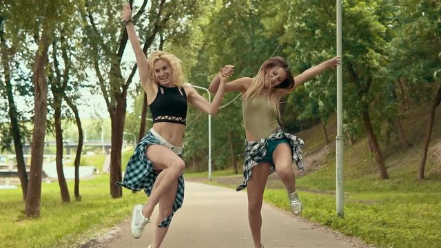 Pretty smiling hipster womans in shorts with shirts jumps in park. Young girls have fun and outdoors in slow motion. Party concept