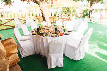 The festive rich round table with white tablecloth and chairs, served with a variety of dishes and drinks, stands on the green grass in the wedding Banquet area against the wooden manor