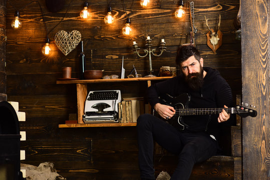 Involved in relaxation. Man with beard holds black electric guitar. Guy in cozy warm atmosphere play relaxing music. Man bearded musician enjoy evening with bass guitar, wooden background
