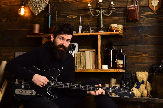 Favourite activity. Man bearded musician enjoy evening with bass guitar, wooden background. Guy in cozy warm atmosphere play relaxing soul music. Man with beard holds black electric guitar