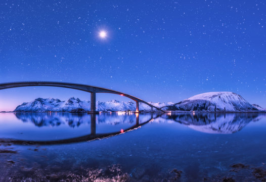Bridge and purple starry sky with beautiful reflection in water. Night landscape with bridge, snowy mountains, sky with full moon and bright stars reflected in sea. Winter in Lofoten islands, Norway - Powered by Adobe