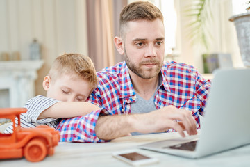 Fototapeta na wymiar Warm toned portrait of cute little boy falling asleep hugging his dad waiting for him to finish working at laptop