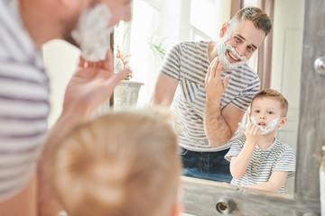 Obraz na płótnie Canvas Warm toned portrait of handsome dad teaching cute little son how to shave, standing against mirror with faces covered in foam in modern bathroom